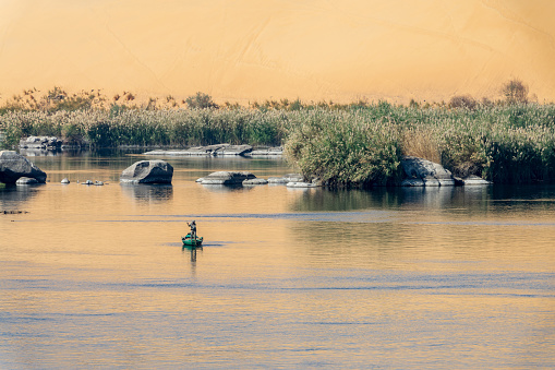 Fisherman in a rowboat on the Nile river at sunet, scenic landscape with rocks in the water and sand dune reflections in Aswan, Egypt