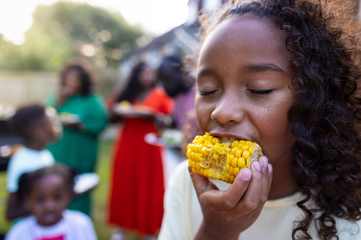Close-up shot of a young girl eating corn on the cob outdoors. She has her eyes closed with other people standing behind her. The people i. the background are out of focus. They are all wearing casual clothing. They're located in Newcastle Upon Tyne, England.\n\nVideos are available similar to this scenario
