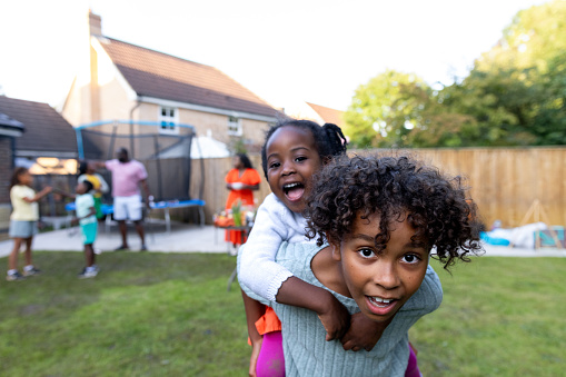 A waist-up shot of a young boy excitably giving a young girl a piggyback and playing in the garden with other family members behind them. They're looking into the camera all wearing casual clothing. They're located in Newcastle Upon Tyne, England.

Videos are available similar to this scenario