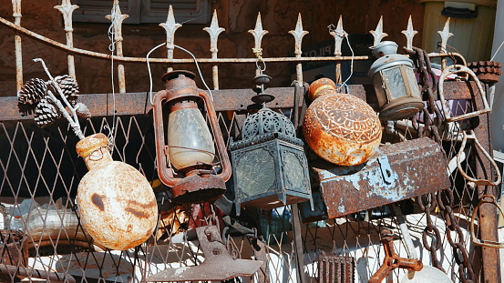 An assortment of aged utensils showcasing signs of rust and peeling paint. Along the fence, lanterns and flags sway, creating a festive ambiance on a cheerful and sunny day. The scene unfolds outdoors.