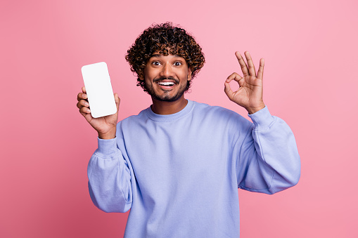 Photo portrait of handsome young male hold white screen device show okey wear trendy blue garment isolated on pink color background.