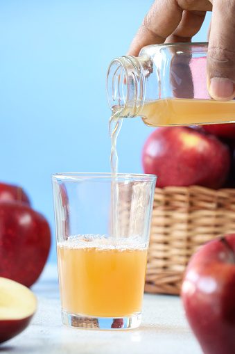 Stock photo showing close-up view of  a wicker basket containing red apples besides a drinking glass into which  apple cider vinegar is being poured from a plastic bottle,  against a blue background. Apple cider vinegar is thought to have several health benefits including weight loss, lower cholesterol, whiten teeth and improve acne. Healthy lifestyle concept.