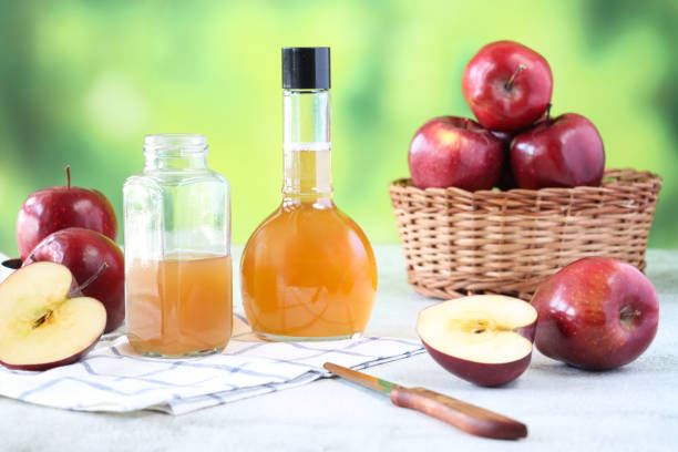 Stock photo showing close-up view of  a wicker basket containing red apples besides two plastic bottles, one open and one closed containing apple cider vinegar,  against a mottled green background. Apple cider vinegar is thought to have several health benefits including weight loss, lower cholesterol, whiten teeth and improve acne. Healthy lifestyle concept.