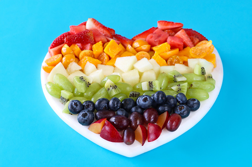 Stock photo showing close-up, elevated view of a plastic, white, heart-shaped chopping board displaying rows of chopped multi-coloured fruits. Rainbow of chopped strawberries, watermelon, mango, physalis (Indian raspberries), honeydew melon, white grape, kiwi, blueberries, plums and red grapes. Healthy eating concept.