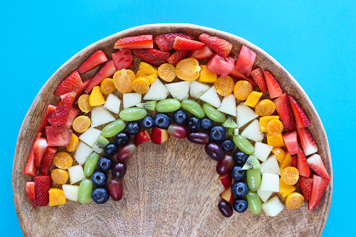 Stock photo showing close-up, elevated view of wooden, chopping board displaying arch of chopped multi-coloured fruits. Rainbow of chopped strawberries, watermelon, mango, physalis (Indian raspberries), honeydew melon, white grape, kiwi, blueberries, plums and red grapes. Healthy eating concept.