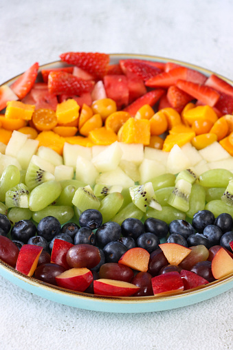 Stock photo showing a close-up, elevated view of plate displaying rows of chopped multi-coloured fruits. Rainbow of chopped strawberries, watermelon, mango, physalis (Indian raspberries), honeydew melon, white grape, kiwi, blueberries, plums and red grapes. Healthy eating concept.