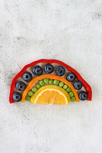 Stock photo showing close-up, elevated view of  arch of chopped multi-coloured fruits and vegetables. Rainbow of strips of red, orange and yellow bell peppers, green peas, blueberries and half slice of orange. Healthy eating concept.