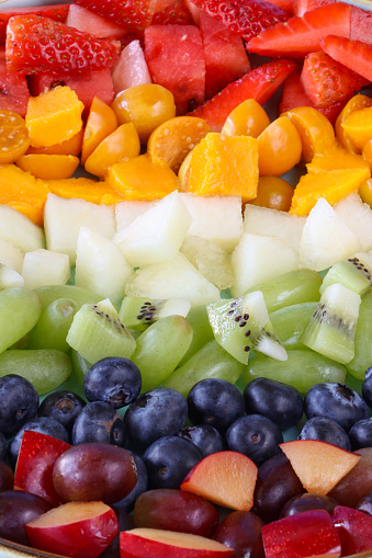 Stock photo showing a close-up, elevated view of a display of chopped multi-coloured fruits. Rainbow of chopped strawberries, watermelon, mango, physalis (Indian raspberries), honeydew melon, white grape, kiwi, blueberries, plums and red grapes. Healthy eating concept.