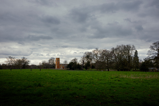A wide angle shot of St Laurences Church, Diddington, with a moody cloudy, sky, winter trees and a field in the foreground.