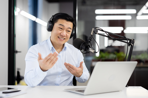 Smiling young Asian man wearing headphones sitting in office in front of desk with microphone and talking on video call on laptop gesturing with hands.