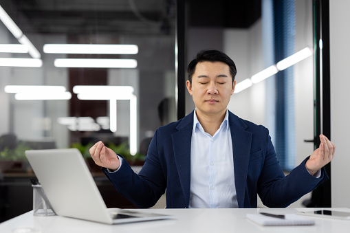 Successful asian boss meditating in lotus position at workplace inside office, man with closed eyes sitting at desk with computer, successful investor banker, dreaming peacefully.