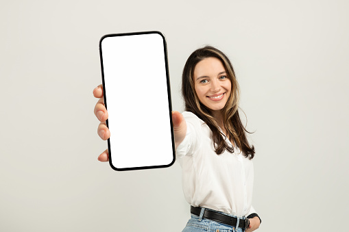 Smiling european young brunette woman in a white blouse presenting a smartphone with a blank screen towards the camera, with a light background providing copy space, studio. Work, business
