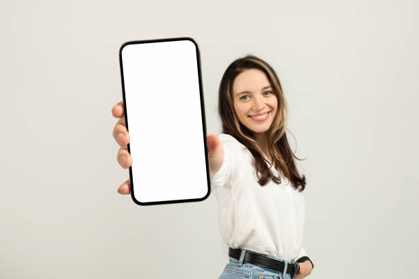 Smiling young brunette woman in a white blouse presenting a smartphone with a blank screen Smiling european young brunette woman in a white blouse presenting a smartphone with a blank screen towards the camera, with a light background providing copy space, studio. Work, business people using mobile phone stock pictures, royalty-free photos & images