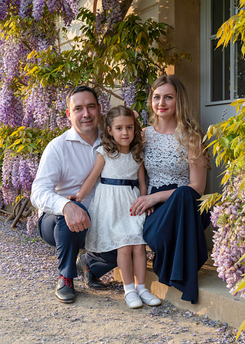 Family portrait. Mom, dad and daughter. Mom and girl are dressed in beautiful dresses, dad is wearing a white shirt. Floral background. Warm, close family relationships