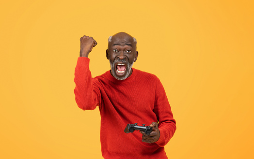 Excited elderly African American man with a beard wearing a red sweater triumphantly raises his fist while holding a video game controller, with a victorious yell on a yellow background