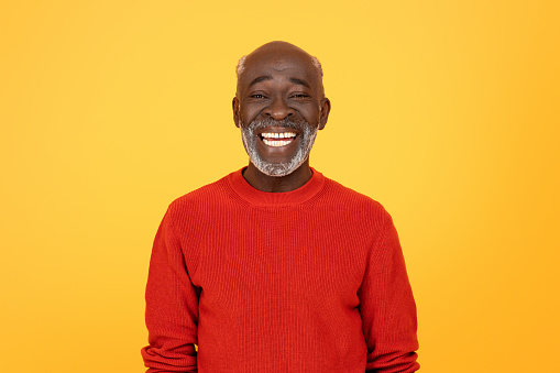 Radiant glad senior african american man with a white beard, sporting a red sweater and a beaming smile, standing confidently against a monochrome yellow studio background