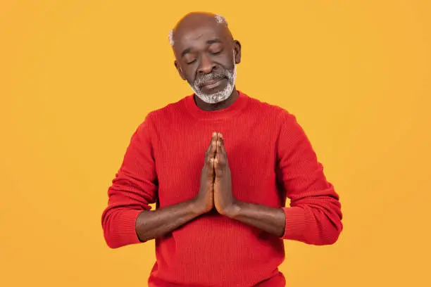 Peaceful senior black man with eyes closed in meditation, hands pressed together in a prayer gesture, wearing a red sweater, embodying calmness and serenity on a yellow background