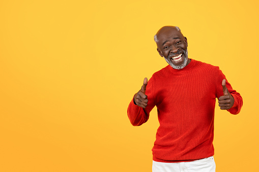 Exultant senior Black man with a radiant smile giving two thumbs up, wearing a festive red sweater against a bright yellow background, symbolizing extreme satisfaction and happiness