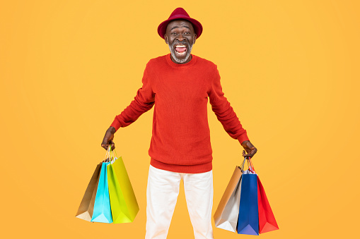 Overjoyed senior african american man in vibrant red attire triumphantly raises colorful shopping bags, capturing the spirit of a successful and enjoyable shopping experience