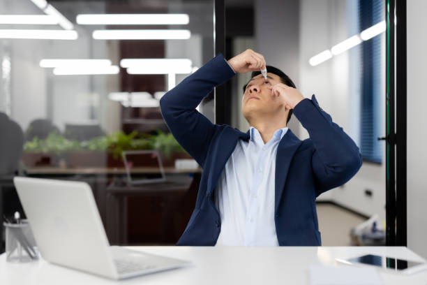 Asian young businessman instills his eyes with medicine drops for inflammation and overfatigue. He sits at his desk in the office in a suit and works overtime Asian young businessman instills his eyes with medicine drops for inflammation and overfatigue. He sits at his desk in the office in a suit and works overtime. soaked suit stock pictures, royalty-free photos & images