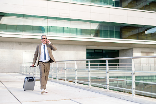 In this outdoor setting, the businessman, engrossed in a phone conversation, walks with his suitcase, portraying the blend of business and mobility.