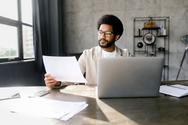istock Young businessman reviewing documents while working on a laptop in an office with an industrial aesthetic 2104167887