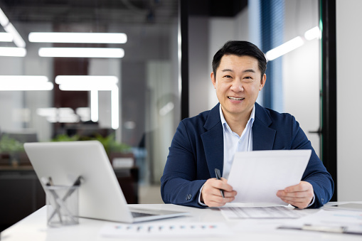 Smiling Asian businessman holding papers, confidently working in a contemporary office environment with a laptop.