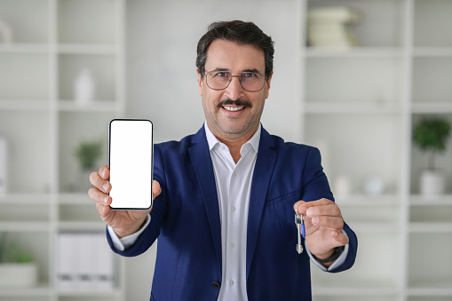 Glad caucasian mature businessman in a smart blue suit holds a mobile phone with a blank screen and a set of keys, posing in an office with a bookshelf backdrop . Work, business recommendation