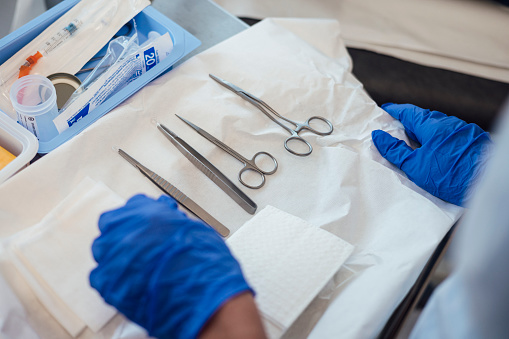 Close-up shot of a doctor holding a tray of medical tools. The tray contains tools such as scissors and tweezers. The doctor is wearing blue rubber gloves whilst holding the tray.