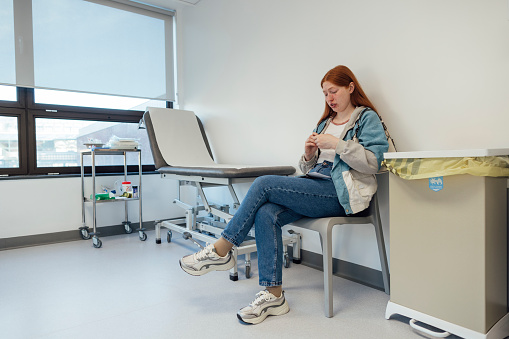 Wide shot of a patient sitting in a hospital room. The woman is playing with her hands while waiting. She is surrounded by an examination bed and medical bins. She has a condition called Behcet's that causes blood vessel inflammation throughout the body.