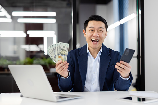 Joyful businessman in suit at office desk laughing with money in hand and a smartphone, expressing success and wealth.