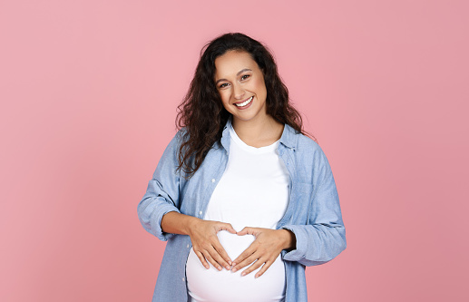Positive expecting millennial European woman hold hands in heart shape on her big tummy, smiling at camera. Happy young pregnant lady posing isolated on pink background, copy space