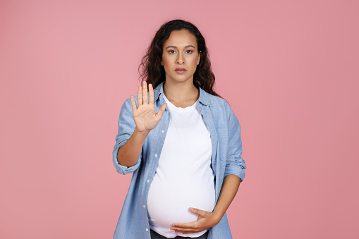 Young regnant woman showing palm rejection gesture while standing isolated over pink background, angry expectant female doing stop sign, demonstrating refusal, copy space