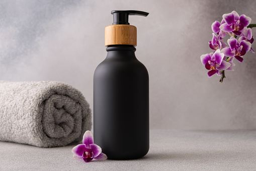 A black matte bottle with a wooden pump cap is centrally placed against a grey backdrop. A rolled grey towel and a vibrant purple orchid flower accentuate the spa-like atmosphere of the image. The scene evokes a sense of luxury and tranquility associated with high-end skincare or wellness products.
