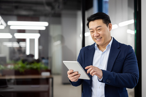 Delighted chinese head of department wearing formal suit and using digital tablet with wide smile. Happy brunette man enjoying modern technology and new opportunities for remote work. Copy space.
