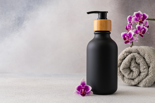 A black matte bottle with a wooden pump cap is centrally placed against a grey backdrop. A rolled grey towel and a vibrant purple orchid flower accentuate the spa-like atmosphere of the image. The scene evokes a sense of luxury and tranquility associated with high-end skincare or wellness products.