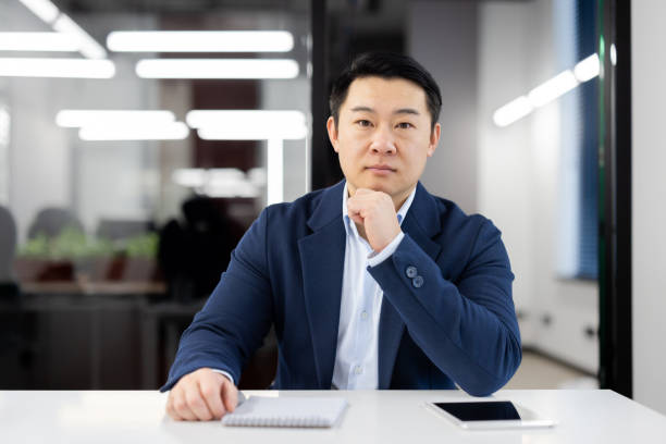 istock Serious asian man in formalwear leaning head on hand while sitting by desk with notebook and tablet. Attentive hr manager getting ready for interview with potential employee and making remarks 2104256564