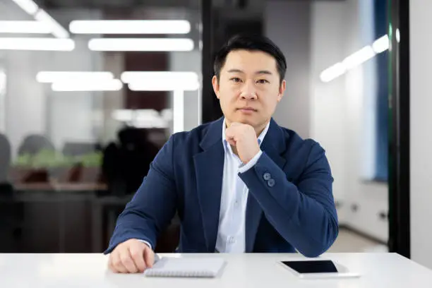 Serious asian man in formalwear leaning head on hand while sitting by desk with notebook and tablet. Attentive hr manager getting ready for interview with potential employee and making remarks.