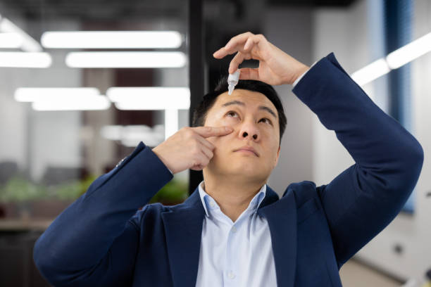 Close-up photo of a young Asian businessman who drips drops of medicine into his eye from overwork and illness. Located in the office behind the workplace Close-up photo of a young Asian businessman who drips drops of medicine into his eye from overwork and illness. Located in the office behind the workplace. soaked suit stock pictures, royalty-free photos & images