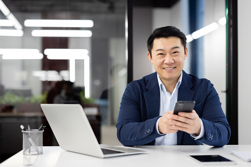 Optimistic asian man holding mobile phone while looking at camera by desktop with pc in modern office interior. Self-employed person having break from work while checking messengers and social media.