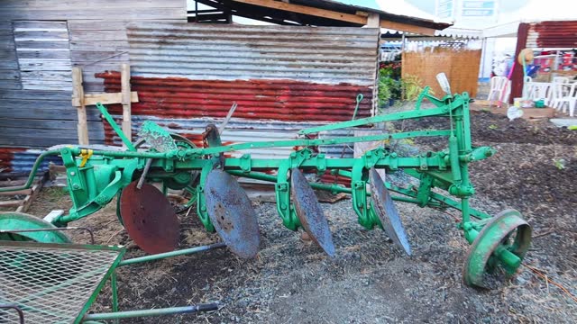 Green bottom plow tool on a soil land by a rusty old metal barn in a rural village in daytime