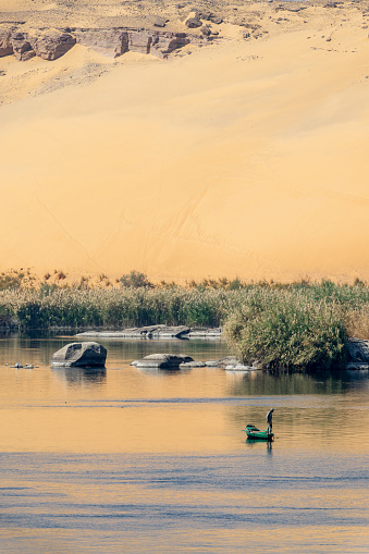 Fisherman in a rowboat on the Nile river at sunet, scenic landscape with rocks in the water and sand dune reflections in Aswan, Egypt