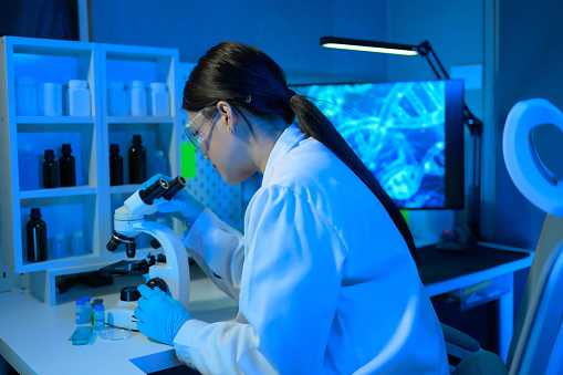 A woman in a lab coat is looking through a microscope. She is wearing a lab coat and gloves