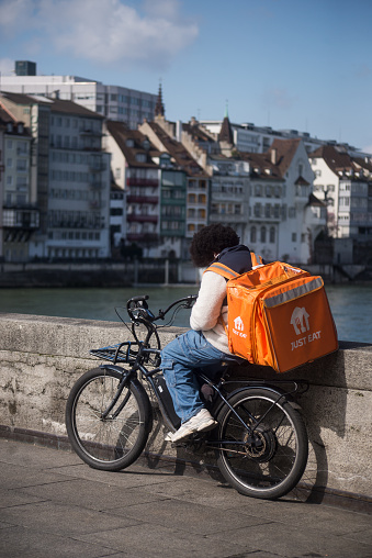 Basel - Switzerland - 21 March 2024 - Portrait of deliver man with orange back pack just eat waiting on bicycle in the street