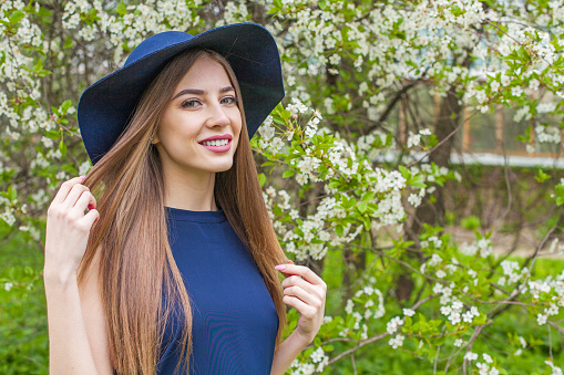 Smiling brunette woman portrait. Beautiful female model with long hair and make-up wearing classic blue hat in spring garden outdoor