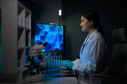 A woman in a lab coat is working on a computer with a monitor displaying DNA. She is wearing gloves and she is focused on her work