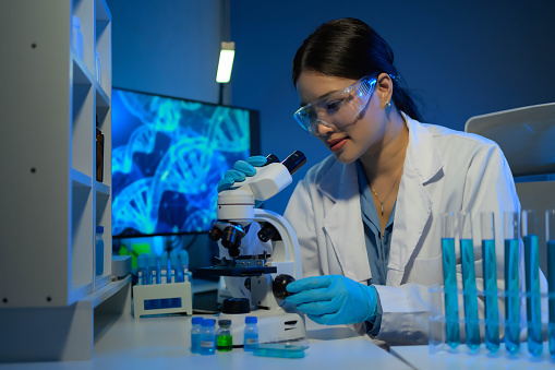 A woman in a lab coat is looking through a microscope at a slide. She is wearing safety goggles and gloves