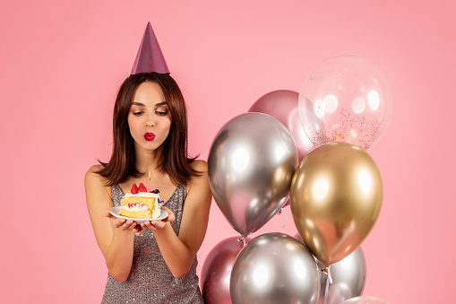 Millennial woman throwing a holiday party or celebration. a pretty woman in a pink dress holds a piece of cake in her hands with candles on the helium balloons. Banner. copy space.