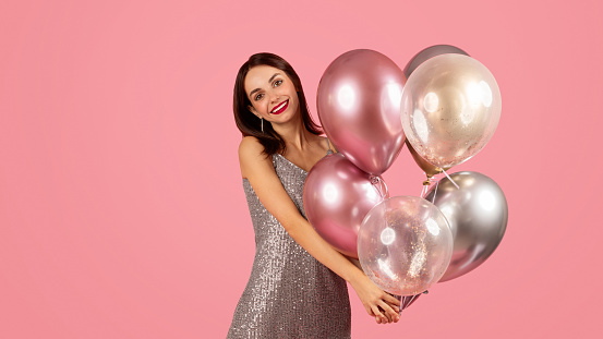 In front of wall with multi colored balloons standing woman in dress and holding heart shaped helium balloon, covering face
