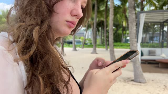 A girl holding a phone rests on the beach in the fresh air Expensive hotel travel ad Chic Luxury beach young woman girl in pareo black swimsuit resting looks at phone uses mobile networks